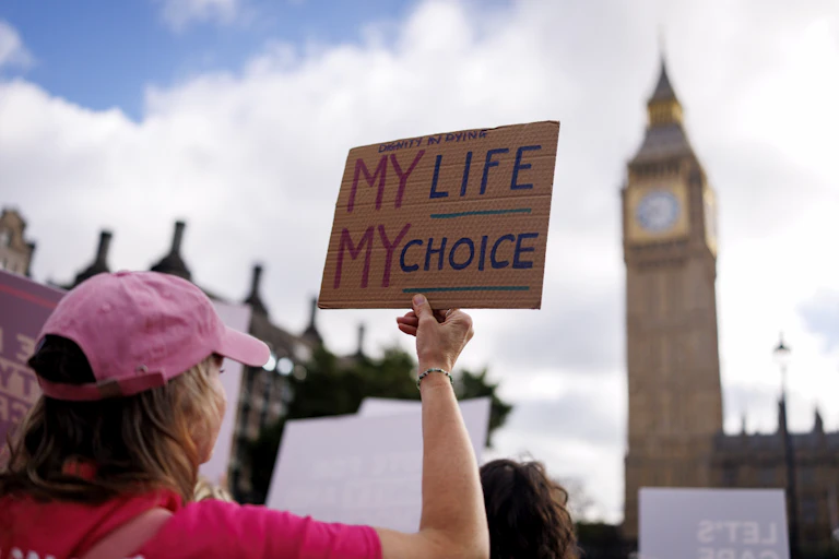pro-assisted dying campaigner in a pink cap in front of big ben holds up a sign saying my life, my choice.