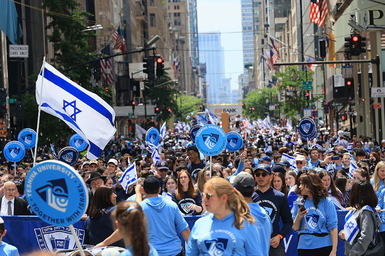 A street of tall buildings, filled with people wearing blue shirts and waving Israeli flags.