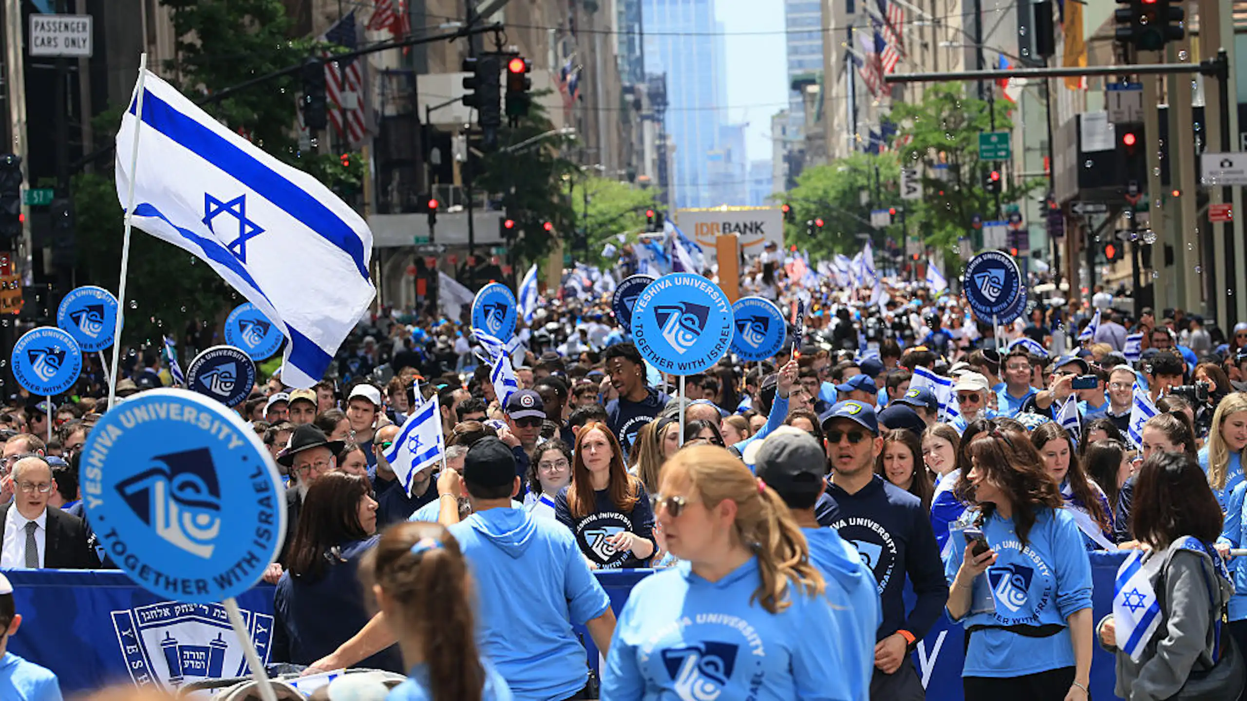 A street of tall buildings, filled with people wearing blue shirts and waving Israeli flags.