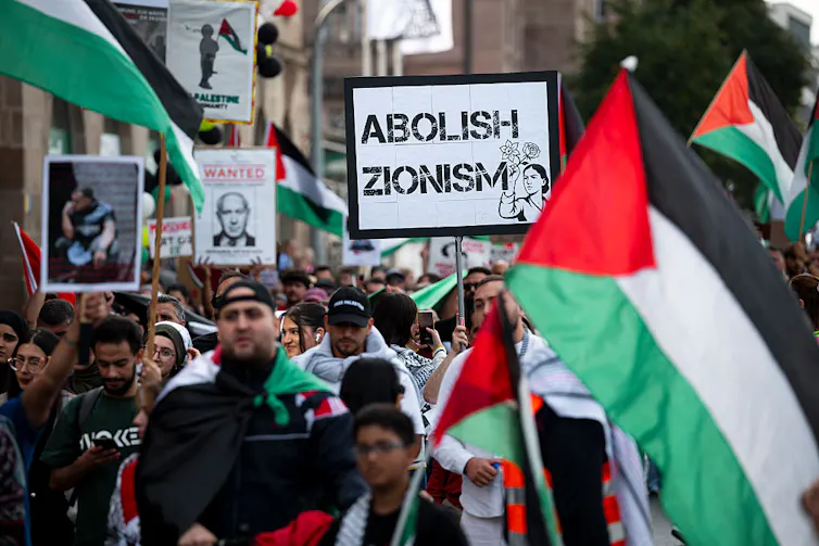 A street full of people marching with Palestinian flags and a sign that says 'Abolish Zionism.'