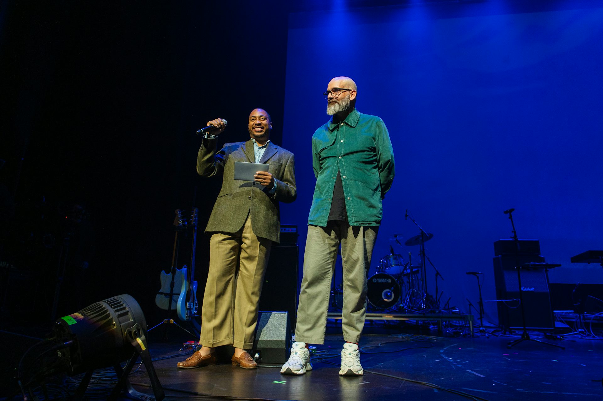 Two men stand on a stage and one holds a microphone.