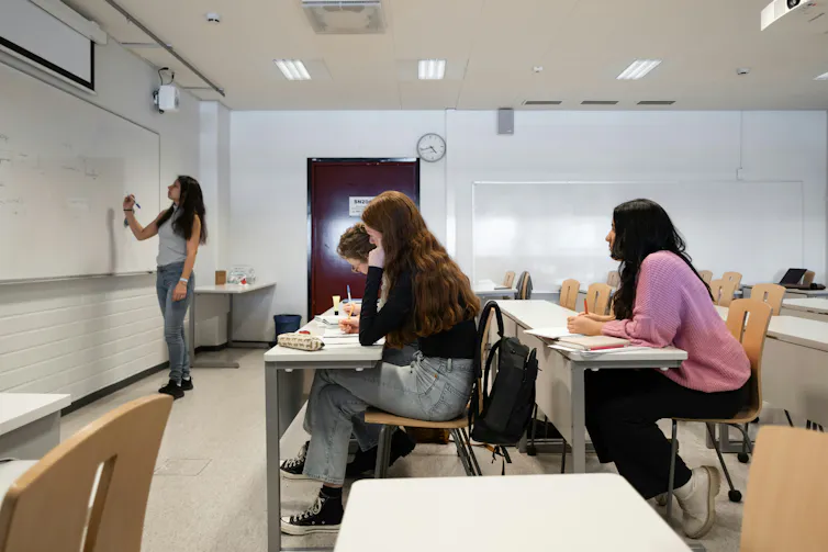 Students sitting in a classroom while an instructor writes on a whiteboard at the front of the room