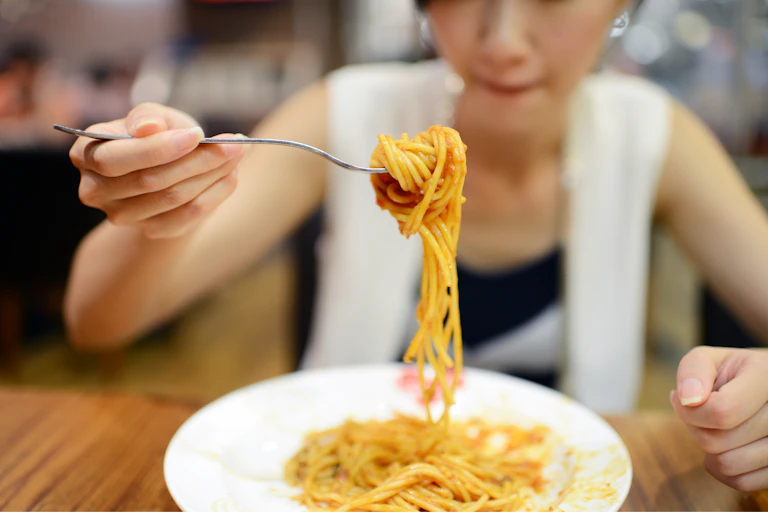 A woman dines on a plate of pasta, with a full fork raised in front of her face.
