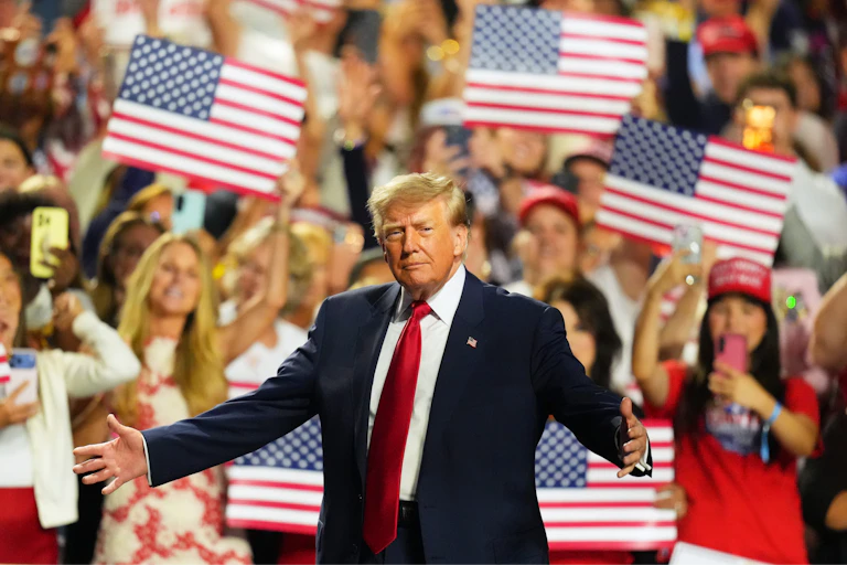 Donald Trump stands in front of a large crowd holding US flags