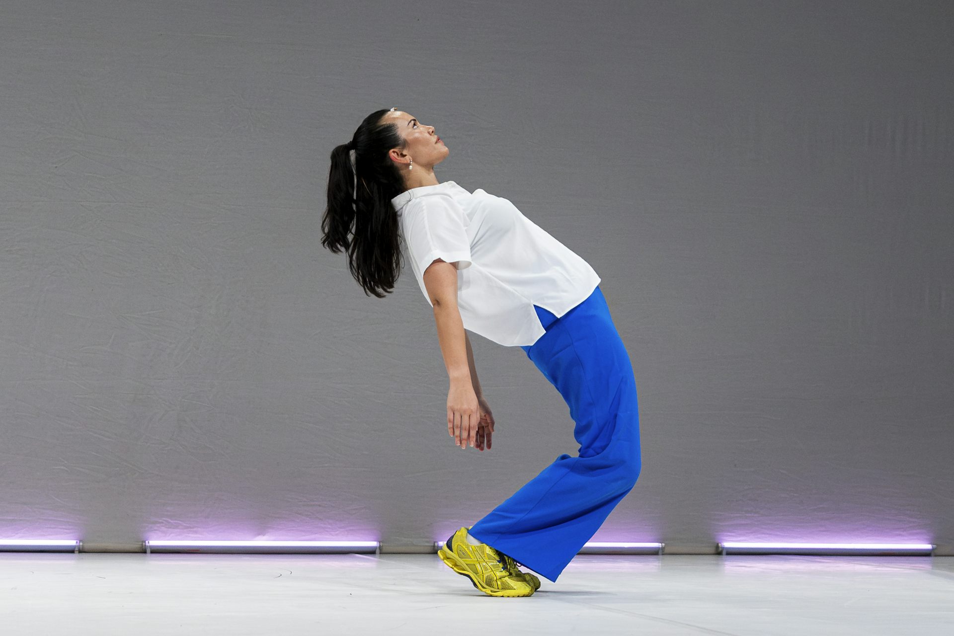 A dancer in blue pants and a white shirt and lime green sneakers arches backwards on a minimalist stage.