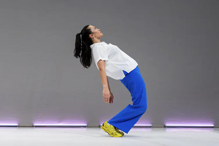 A dancer in blue pants and a white shirt and lime green sneakers arches backwards on a minimalist stage.