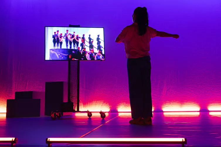 A dancer stands on a pink-lit stage watching a video screen showing footage of dancers in formation.
