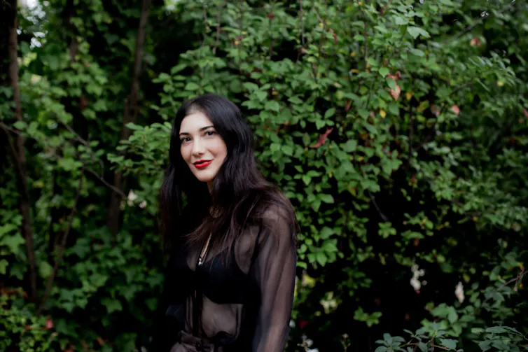 The author, Laura Elizabeth Woollett, a beautiful woman with long, dark hair and red lipstick in front of a hedge.