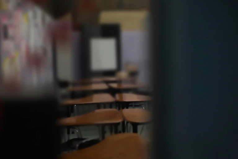 A series of wooden desks and chairs are lined up in a dark room that is partially obscured.