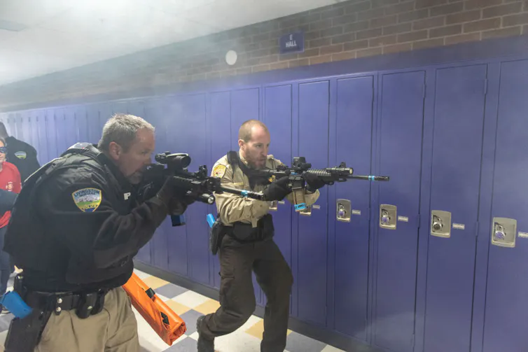 Two men in police uniform hold up large guns near their faces near a row of blue lockers.