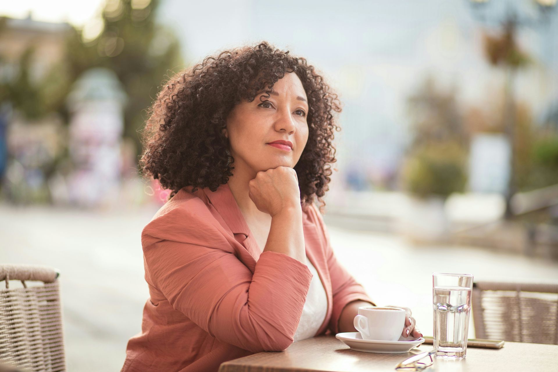 A woman sitting outdoors at a table with a cup of coffee, looking to her right with a slight smile on her face.