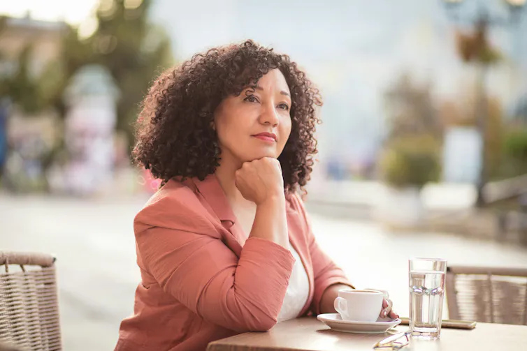 A woman sitting outdoors at a table with a cup of coffee, looking to her right with a slight smile on her face.