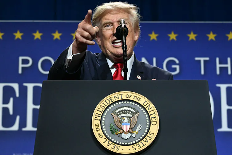A man in suit and tie points to a crowd from an onstage podium.