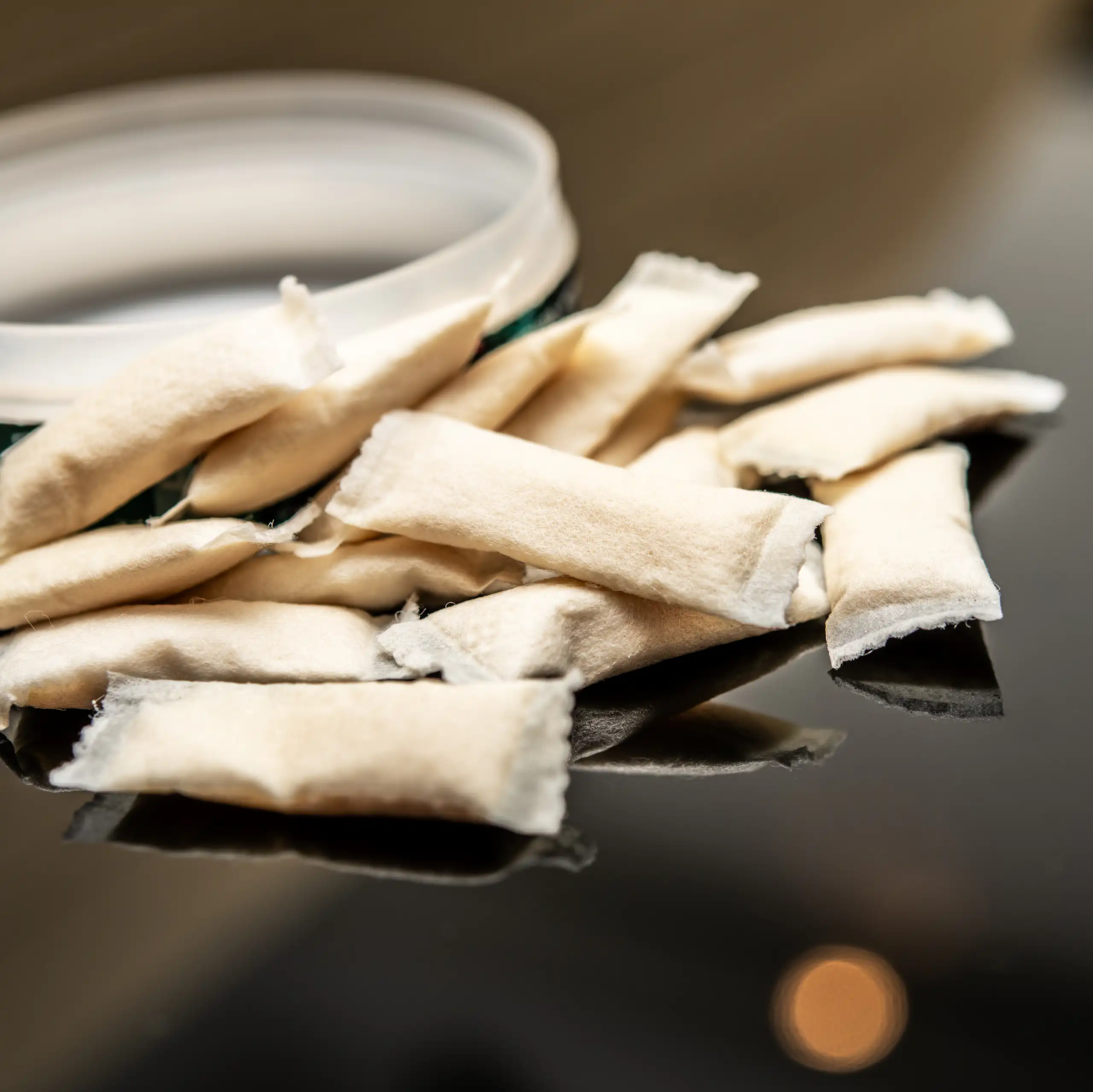 More than a dozen white smokeless tobacco pouches scattered on a reflective black surface next to an open round container in indoor lighting.