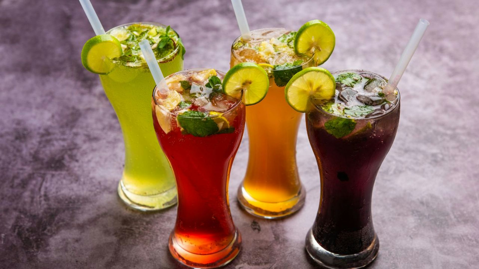 Four tall cocktail glasses with colorful drinks. In order from right: green, red, orange, and purple. Classes also contain mint leaves, ice, lemons and limes.