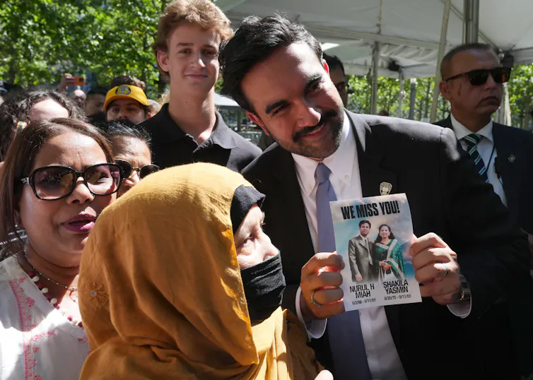 Zohran Mamdani, mayor of New York, in a suit and tie, with a crowd, including a veiled woman.