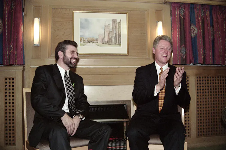 Two men. in suits sit in front of a painting.