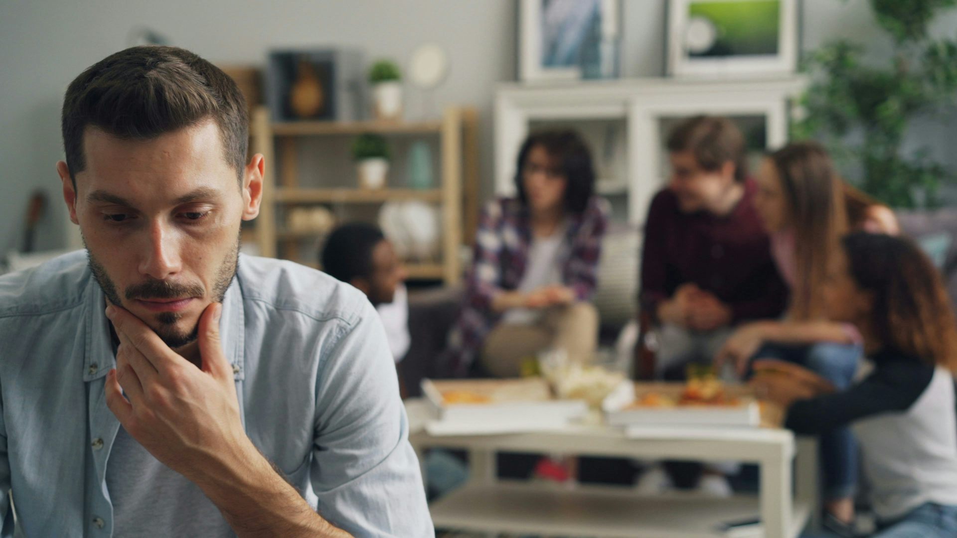 A closeup of a young man thinking while a group of young people sitting around a table in the background are having a conversation.