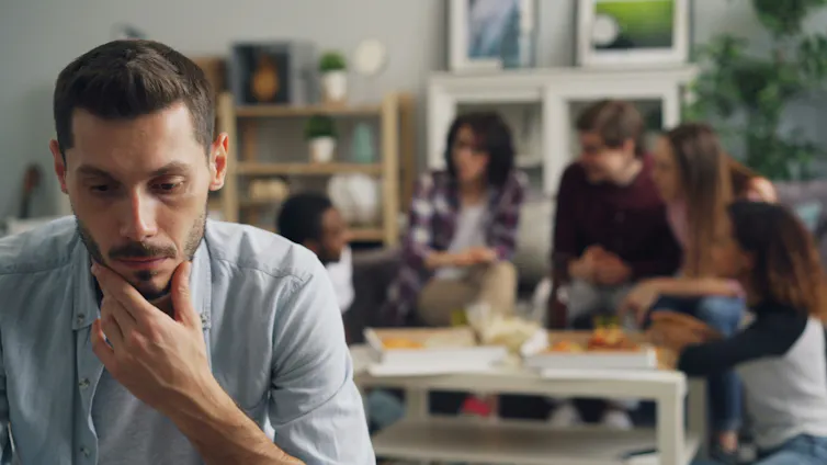 A closeup of a young man thinking while a group of young people sitting around a table in the background are having a conversation.