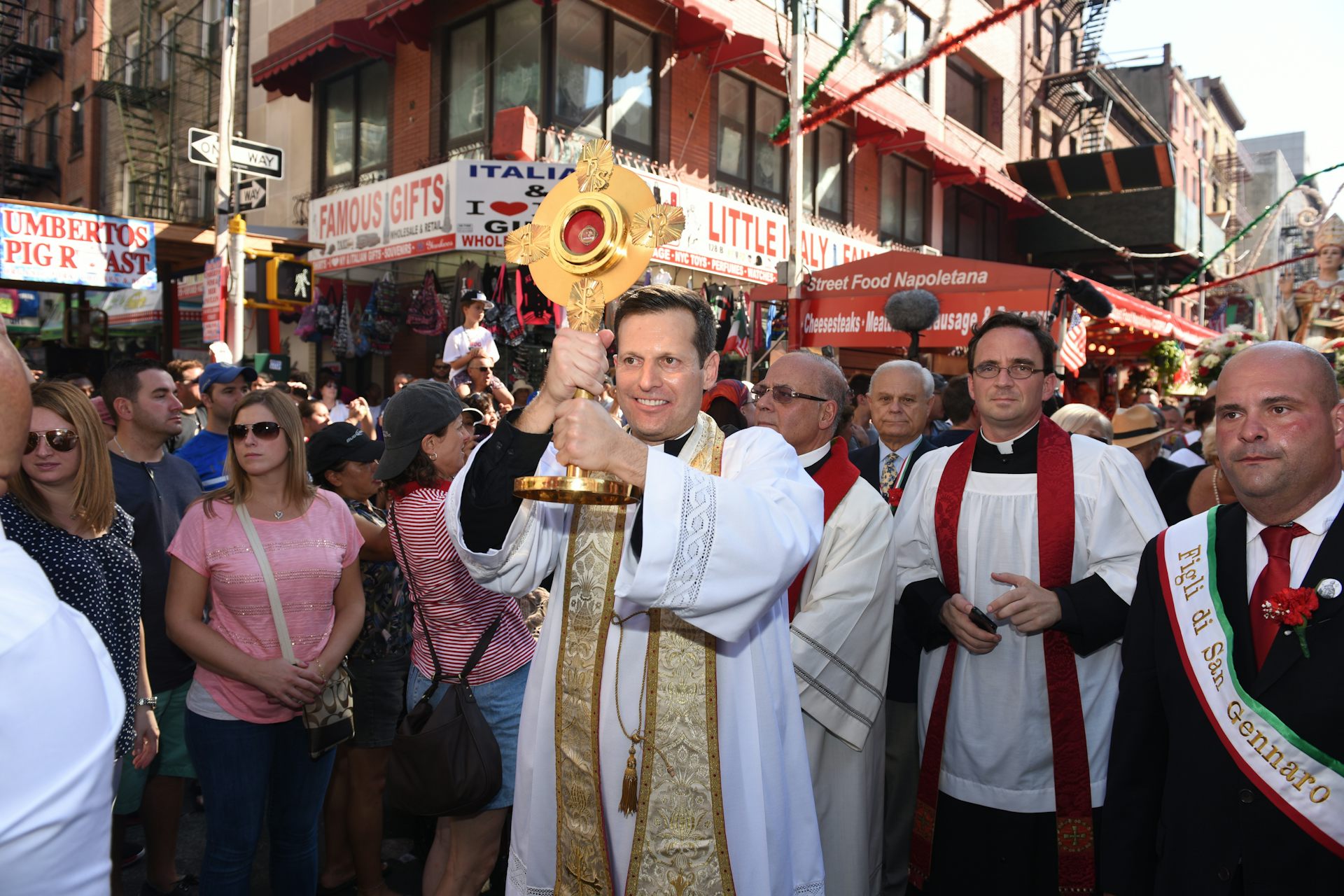 A group of clerics in religious garb, with one holding a large, gold cross, march along a city street as people look on.