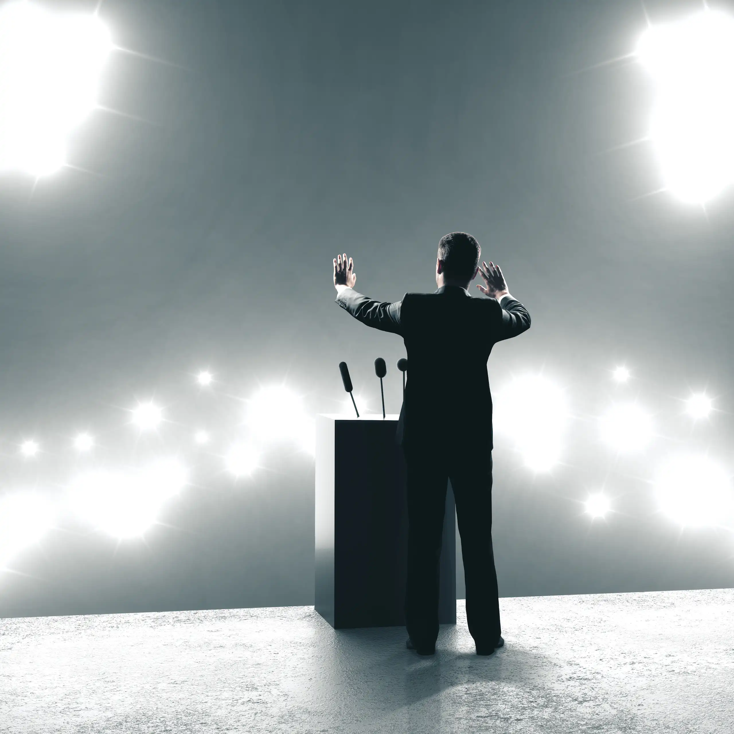 man in a suit standing in the spotlight on a stage with his arms raised to the crowd.