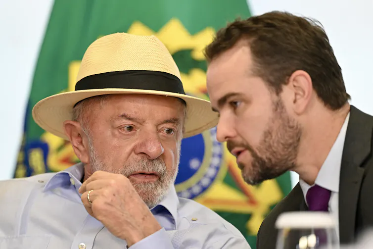 An older man in a straw hat confers with a younger man with a beard with a Brazilian flag in the background.