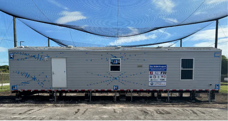 A mobile home stands on a field with a net over it to catch flying debris ahead of a test in the Wall of Wind.