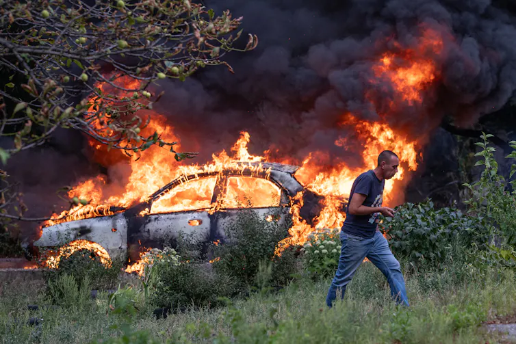 A man wearing jeans and a t-shirt runs through a field away from a car that is on fire, with flames leaping from it