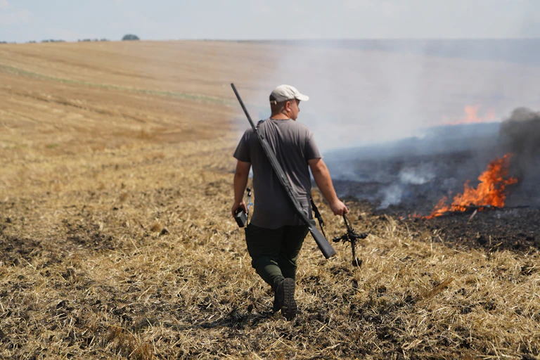 A man walks through a harvested corn field with a rifle slung over this back and a destroyed drone hanging from his hand, as the field burns to his right