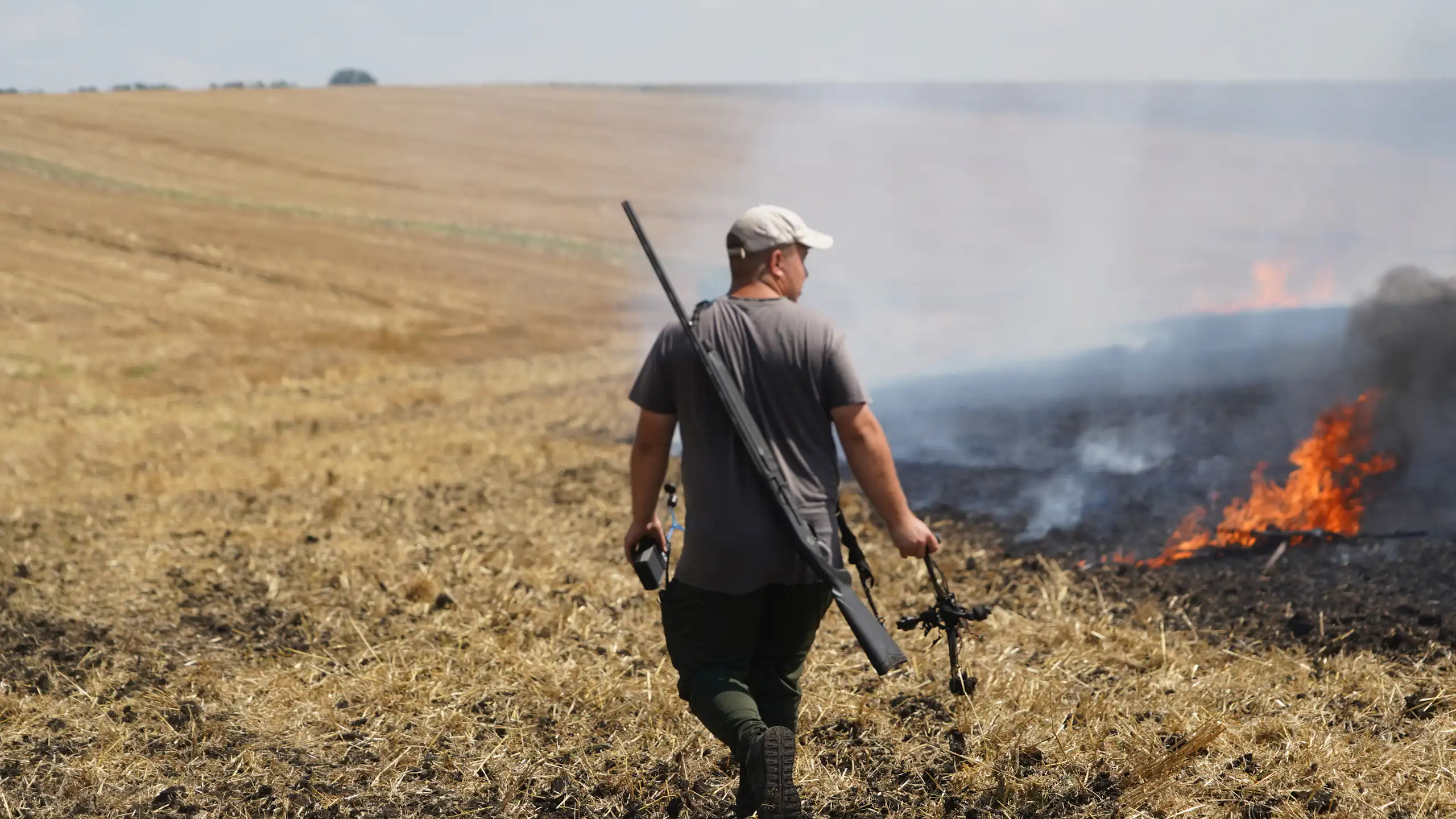 A man walks through a harvested corn field with a rifle slung over this back and a destroyed drone hanging from his hand, as the field burns to his right