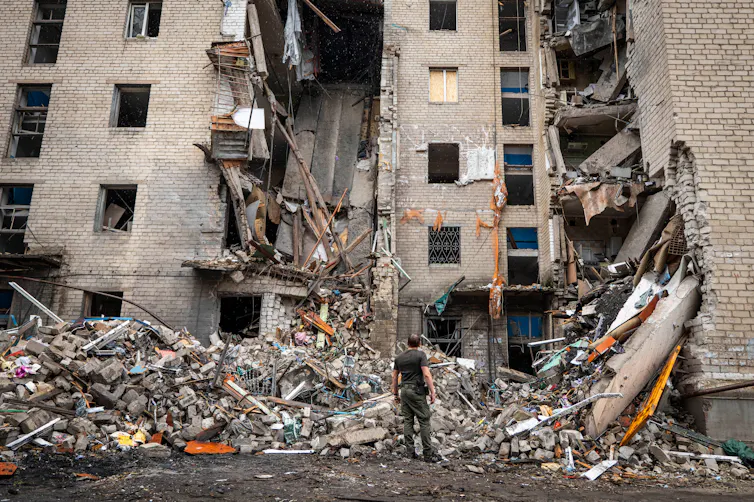 A tall residential apartment building partially damaged with a chunk missing from the middle and a large pile of debris at the bottom