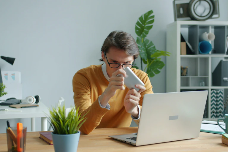Un jeune homme assis devant son ordinateur regarde son téléphone.