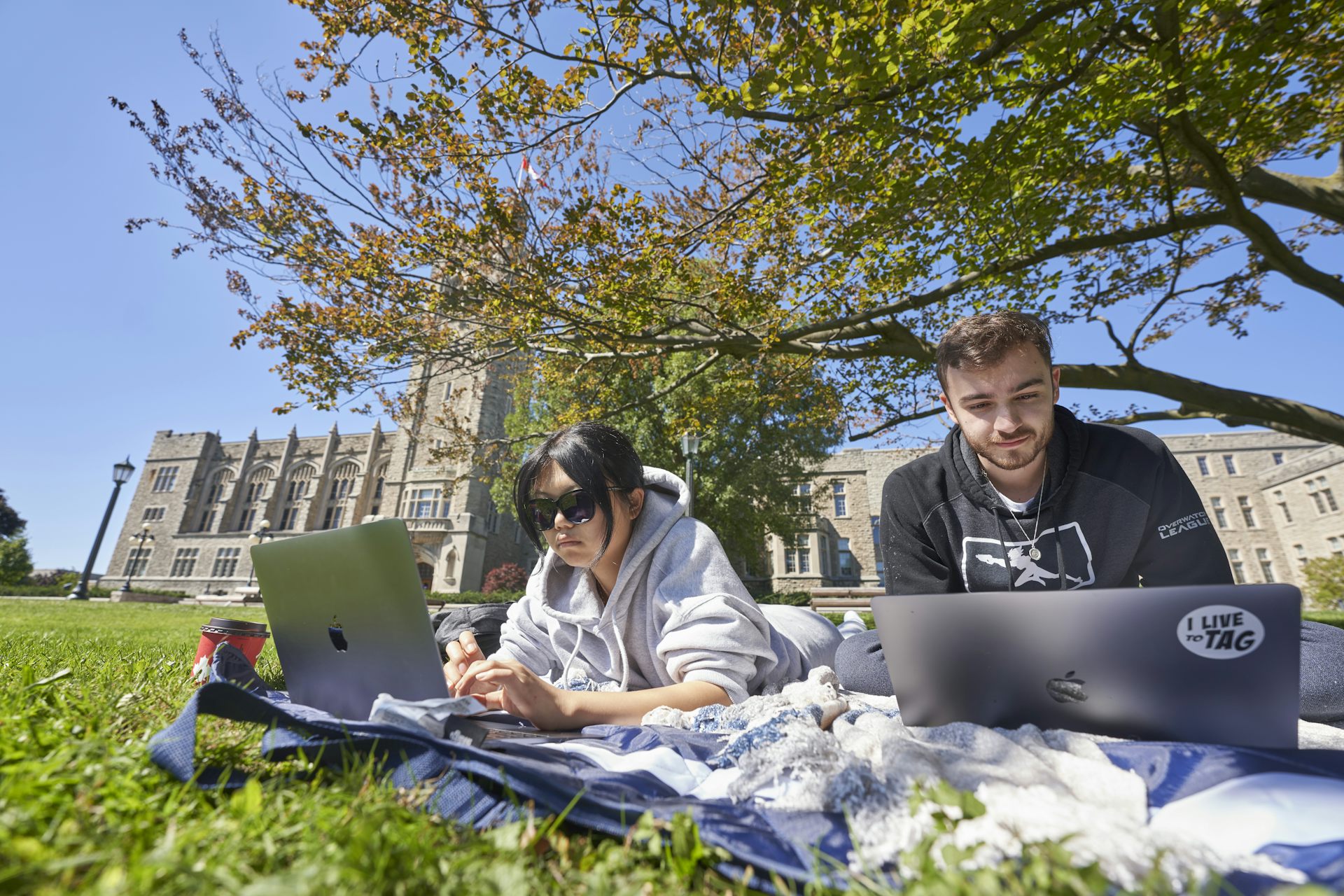 Two university students lie on their stomachs on a blanket in the shade of a tree while on laptops