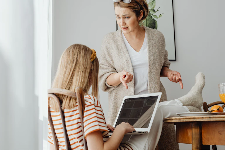 Un enfant a les pieds sur la table et regarde un ordinateur portable, tandis qu’un parent pointe ses pieds du doigt.