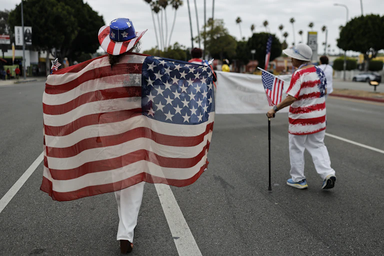 Two people wearing and holding American flags walk down the middle of a street in a parade, facing away from the camera.