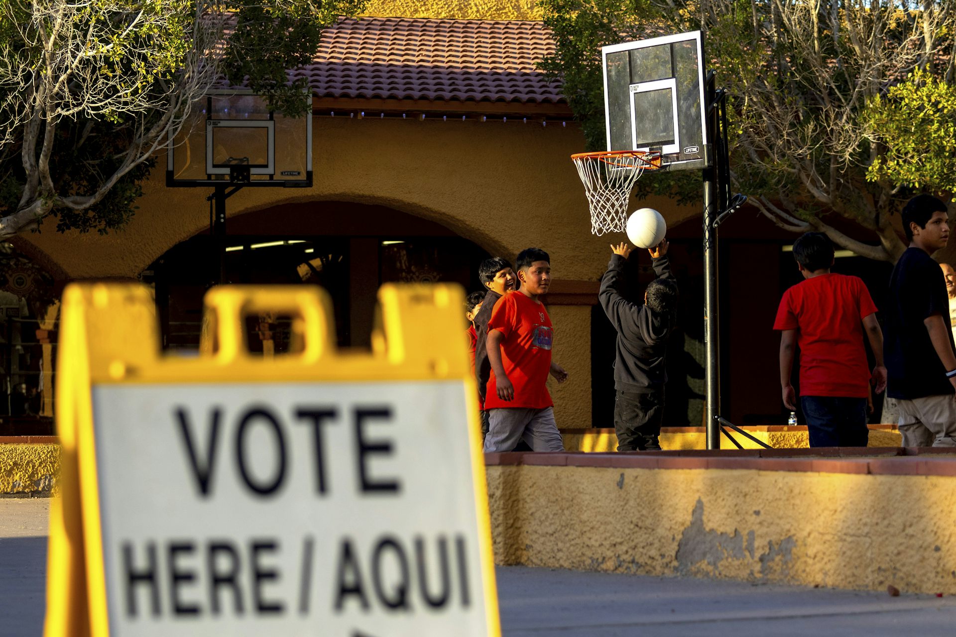 A 'Vote here / Aqui' sign stands in the foreground as young people play basketball outside a building in the background.