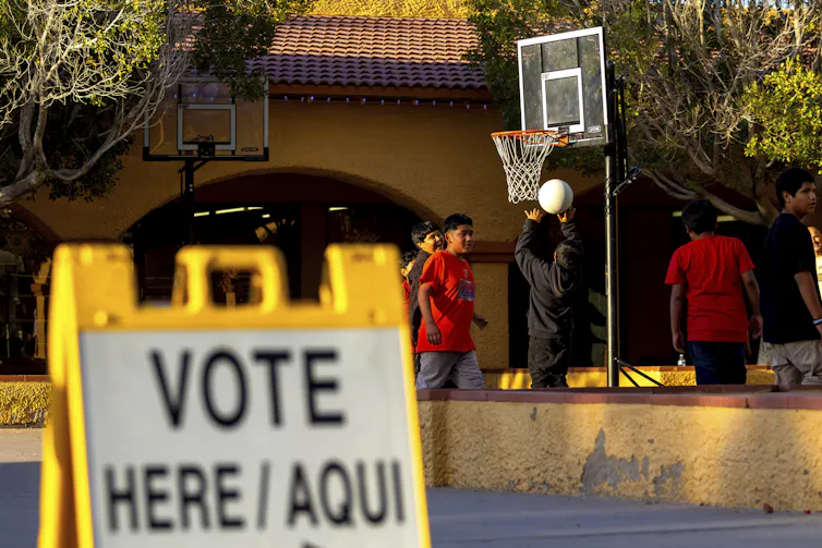 A 'Vote here / Aqui' sign stands in the foreground as young people play basketball outside a building in the background.