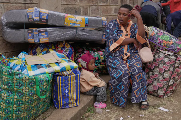 A woman in a colorful gown sits on a pavement with her belongings and child while making a phone call.