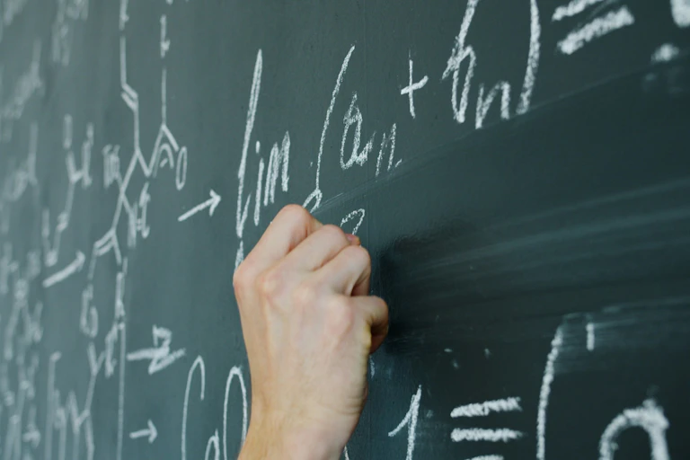 A human hand writes math equations in chalk on a classroom chalkboard.
