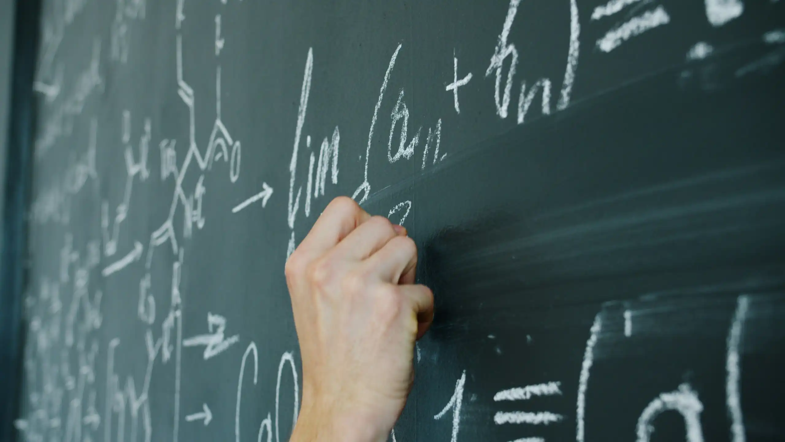 A human hand writes math equations in chalk on a classroom chalkboard.