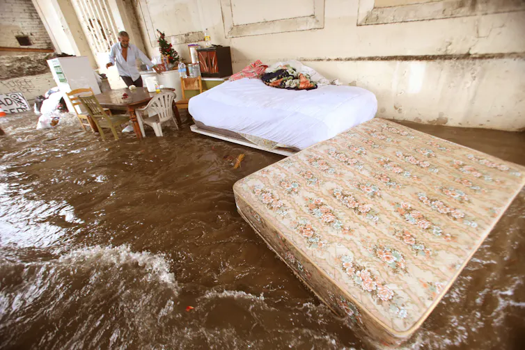 A man walks carefully through water as two mattress float away in a rush of water under a bridge.
