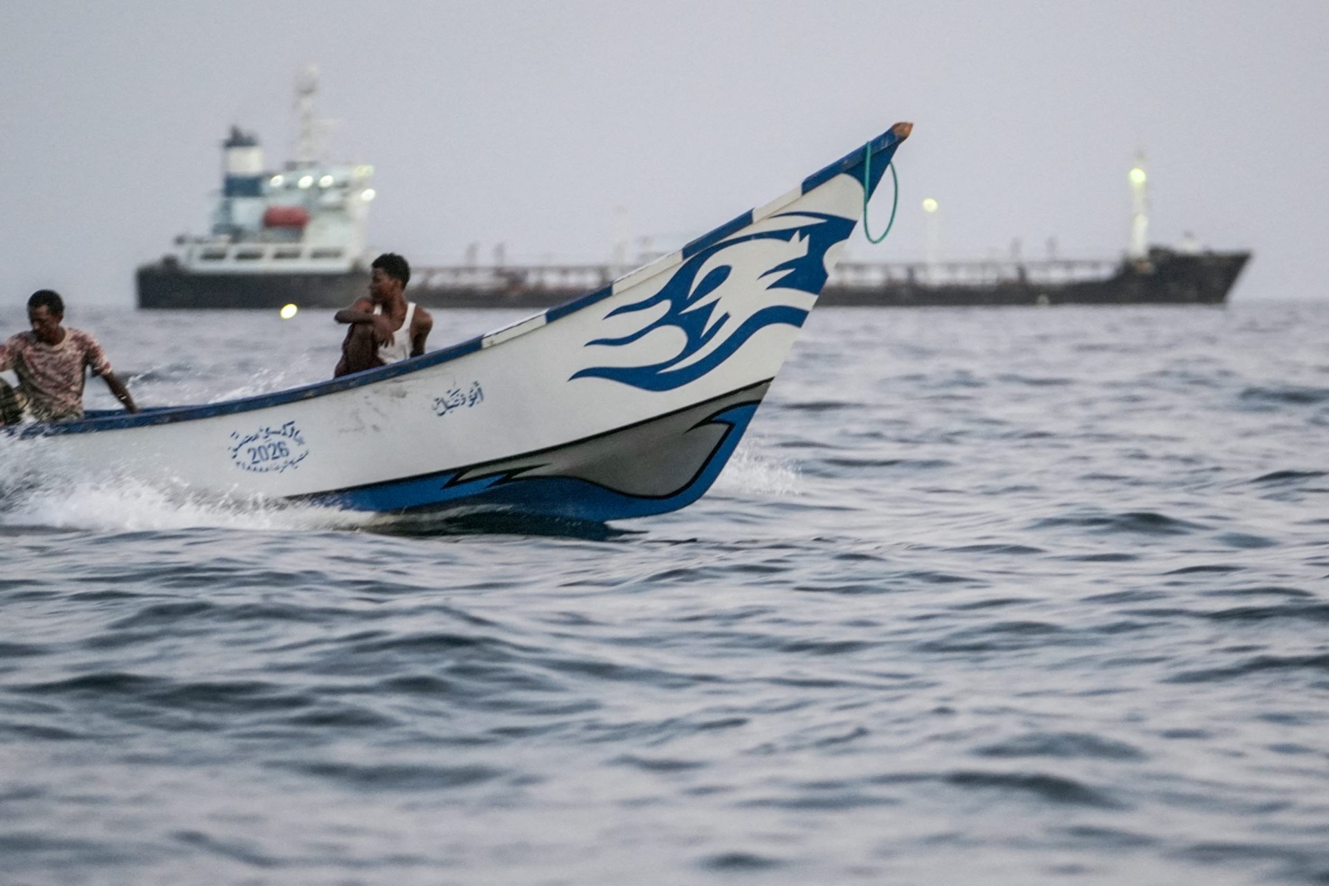 A man on a fishing vessels with a commercial tanker seen in the background.