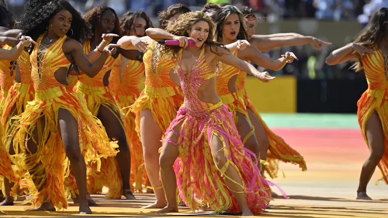 Una mujer canta y baila rodeada de bailarines en medio de un estadio.