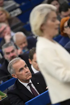 A man with short grey hair in a suit sits behind a blond woman listening as she speaks.