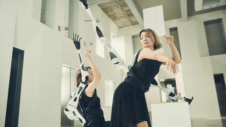 Two people wearing Jizai Arms, a pair of additional robotic arms attached to the back