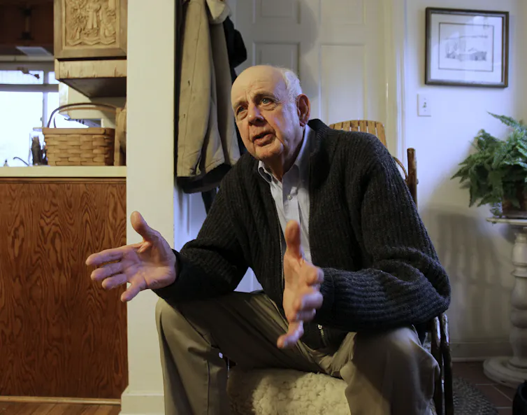 An elderly man sitting inside leans forward and gestures with his hands as he speaks.