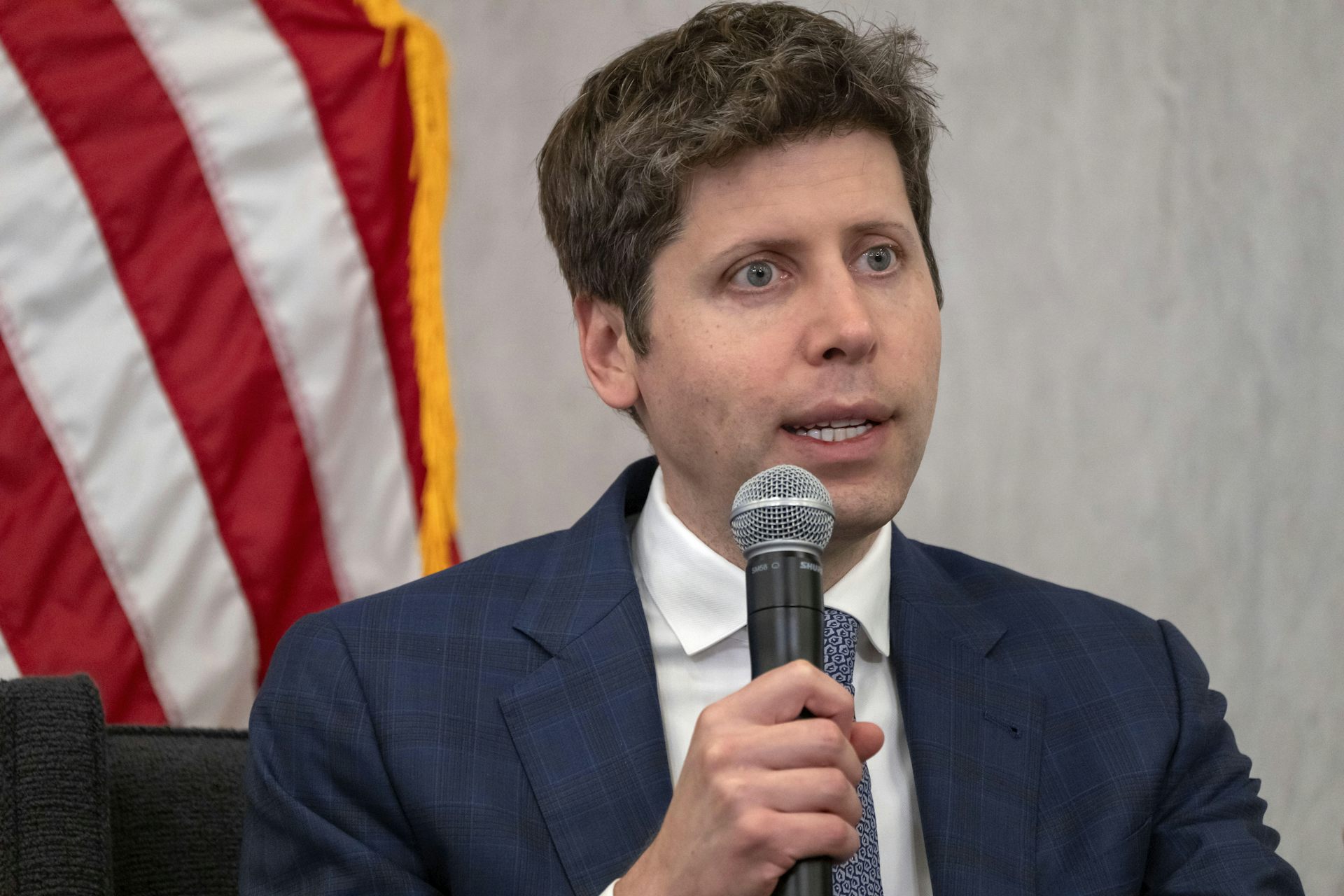 A middle-aged white man in a suit speaks into a microphone while seated in front of an American flag