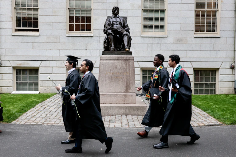 Four young men in graudation gowns walk past a statue on a pedestal.