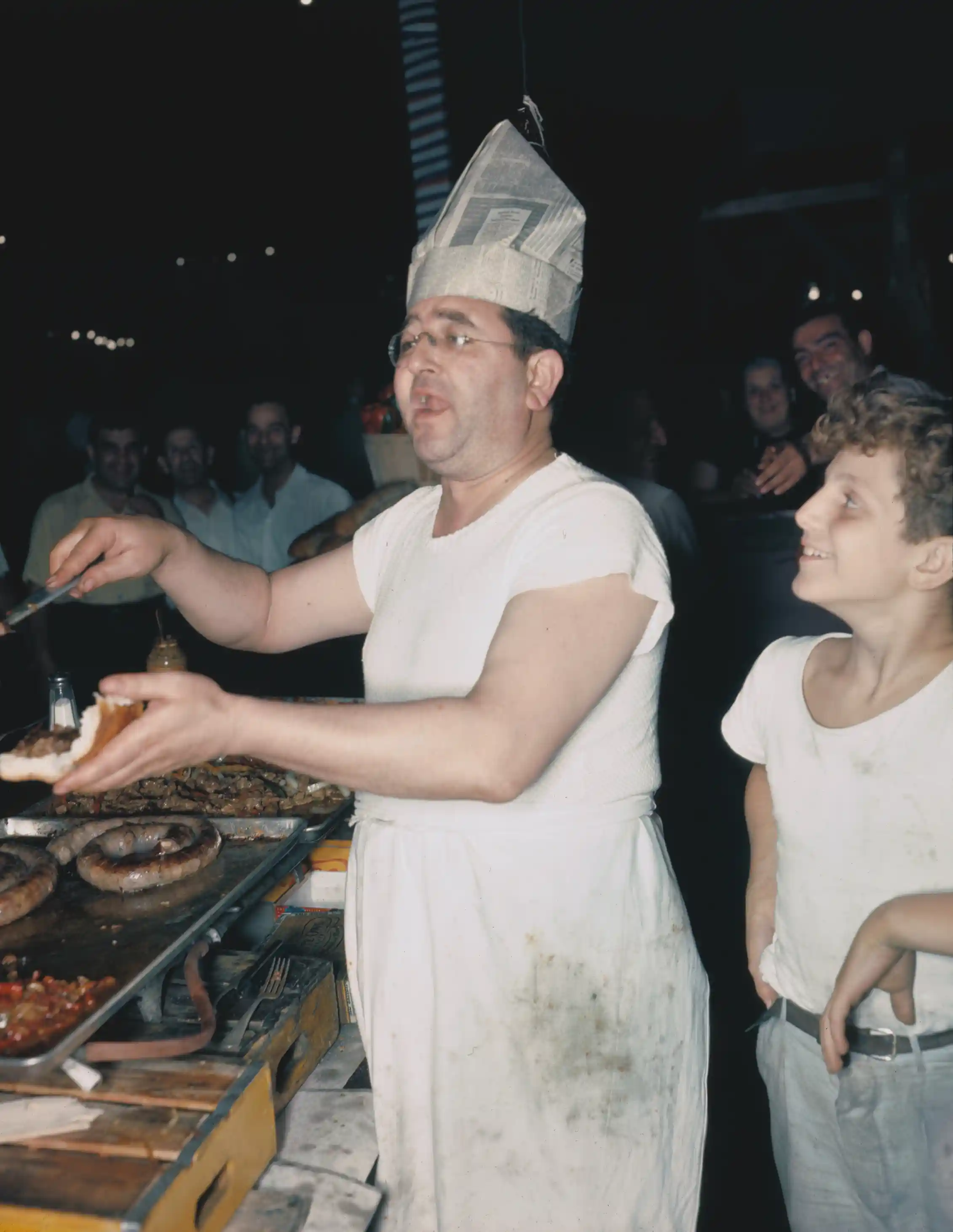 Middle-aged man wearing a greasy, white outfit and a hat made from a newspaper shouts while placing onions, peppers and sausage on a sub roll as a smiling boy l