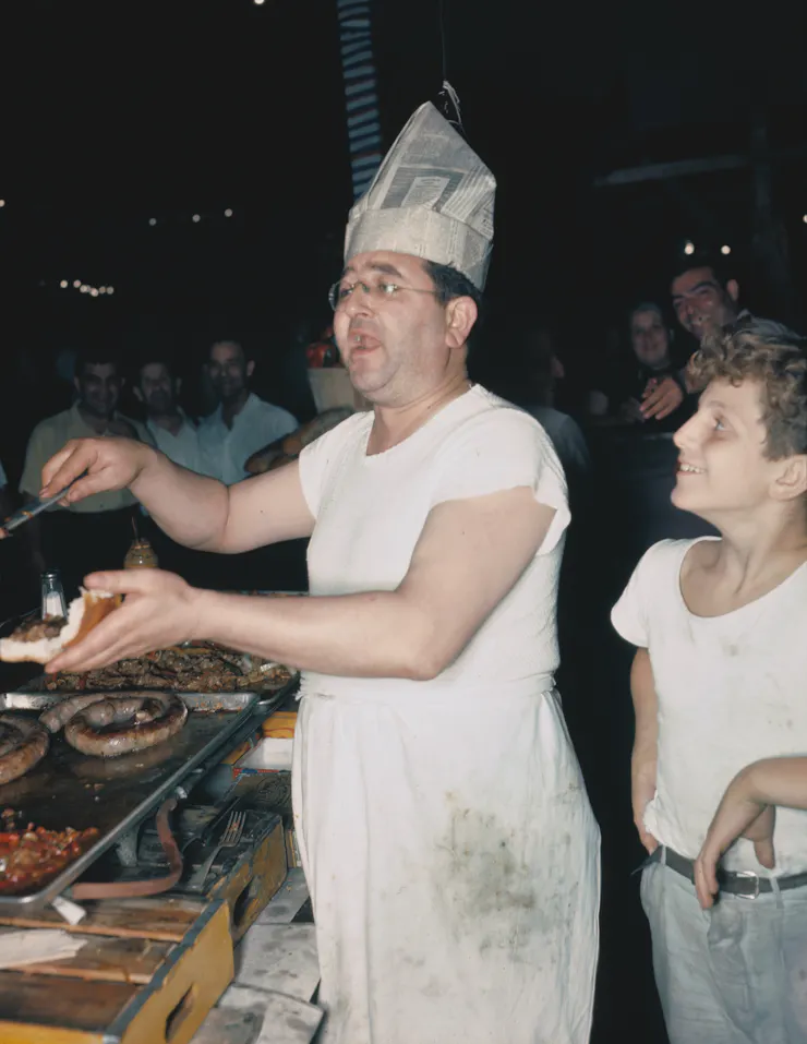 Middle-aged man wearing a greasy, white outfit and a hat made from a newspaper shouts while placing onions, peppers and sausage on a sub roll as a smiling boy looks on.